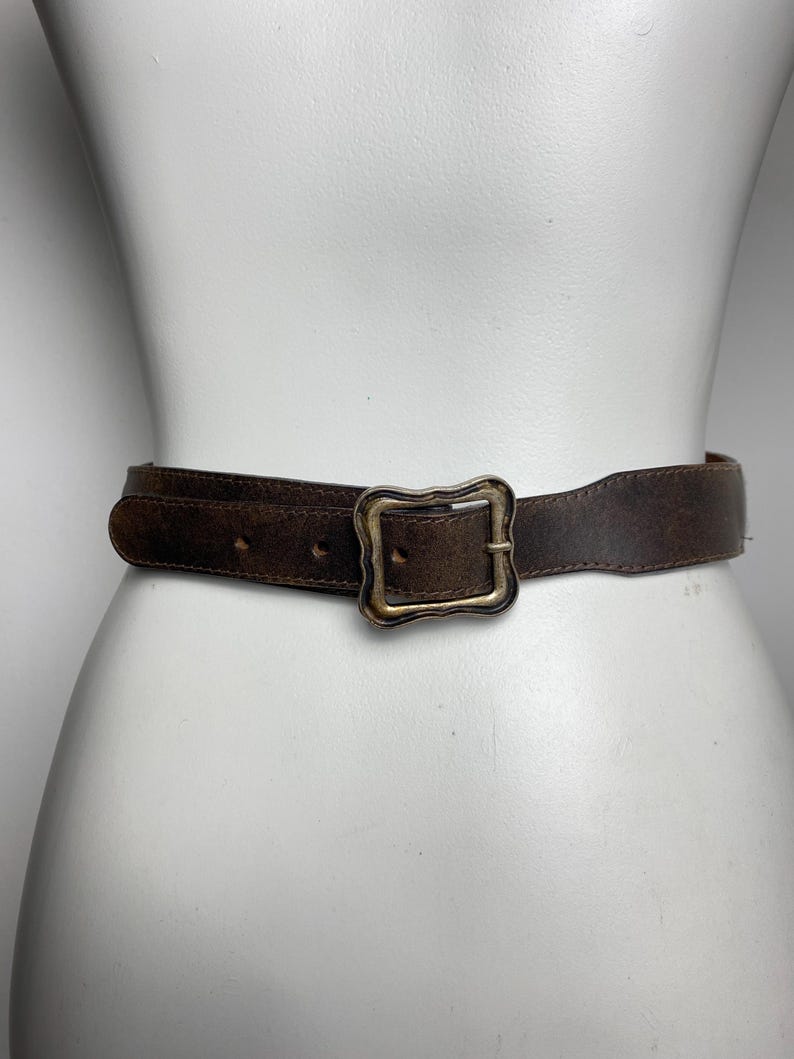 May include: A dark brown leather belt featuring a weathered, distressed texture and a rectangular brass-toned metal buckle with a wavy, decorative edge. The belt has visible stitching along the edges and multiple punched holes for adjustment.