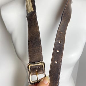 May include: A dark brown distressed leather belt featuring a textured surface and a rectangular brass-toned buckle with scalloped edges. The belt includes a section of light tan fibrous material near the top and has four visible adjustment holes.