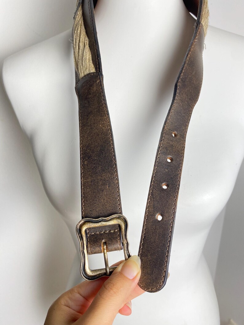 May include: A dark brown distressed leather belt featuring a textured surface and a rectangular brass-toned buckle with scalloped edges. The belt includes a section of light tan fibrous material near the top and has four visible adjustment holes.