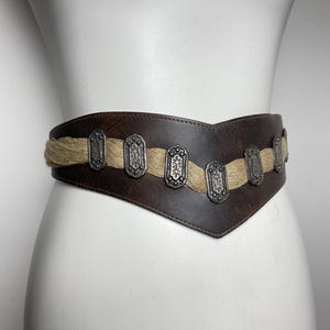 May include: A wide, dark brown leather belt featuring a V-shaped front design. A horizontal strip of natural-colored jute fiber runs across the center, accented by a series of rectangular, silver-toned metal plates with intricate embossed patterns.