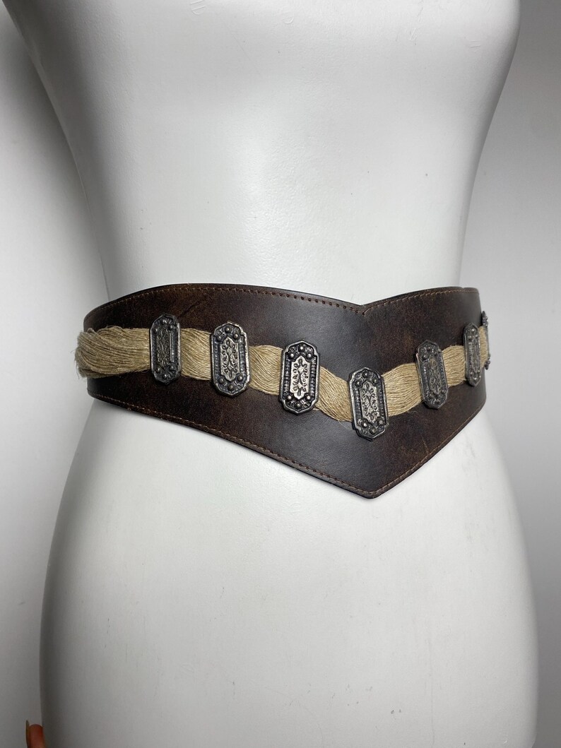May include: A wide, dark brown leather belt featuring a V-shaped front design. A horizontal strip of natural-colored jute fiber runs across the center, accented by a series of rectangular, silver-toned metal plates with intricate embossed patterns.