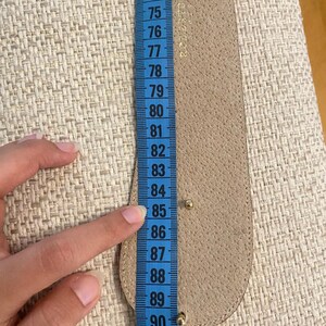 May include: A beige leather belt is measured with a blue measuring tape. The belt is approximately 33 inches long. The measuring tape shows the belt's length in inches, with numbers from 72 to 92.