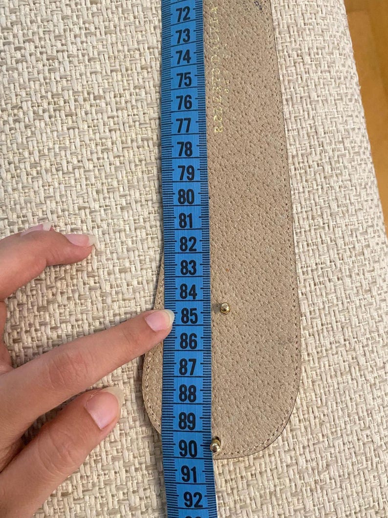 May include: A beige leather belt is measured with a blue measuring tape. The belt is approximately 33 inches long. The measuring tape shows the belt's length in inches, with numbers from 72 to 92.