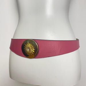 May include: A pink leather belt with a gold-colored sunburst buckle. The belt is wide and tapers towards the back. The buckle has a central jewel. The belt is displayed on a white mannequin.