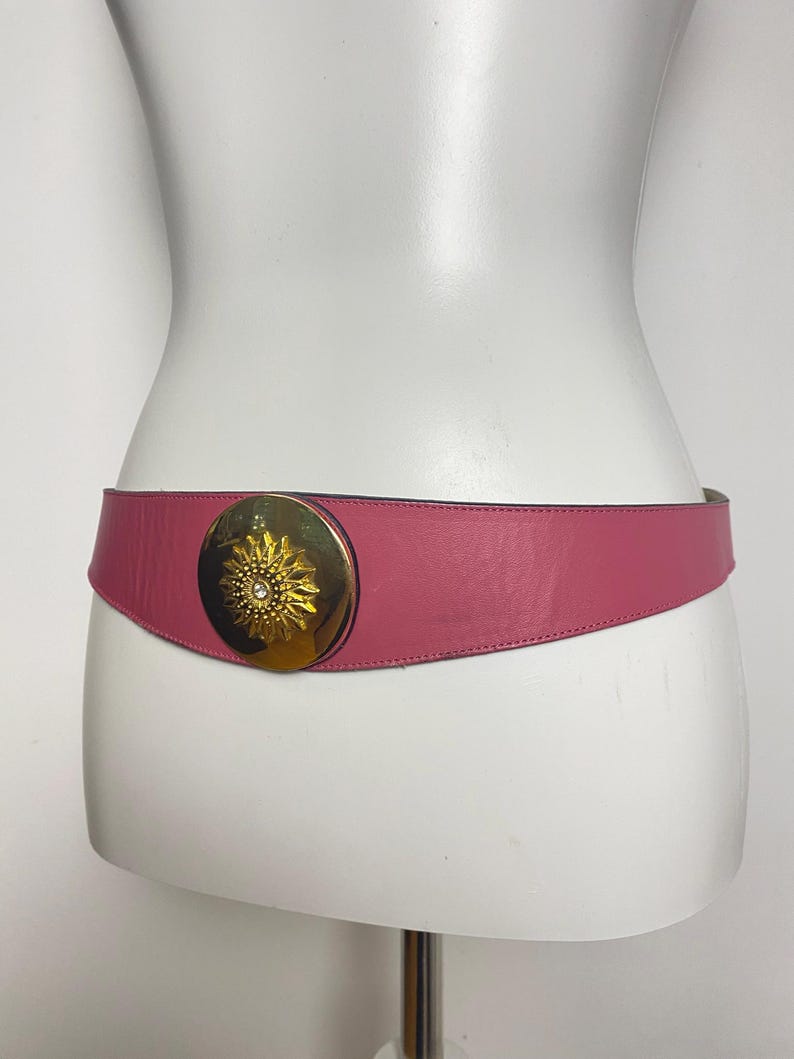 May include: A pink leather belt with a gold-colored sunburst buckle. The belt is wide and tapers towards the back. The buckle has a central jewel. The belt is displayed on a white mannequin.