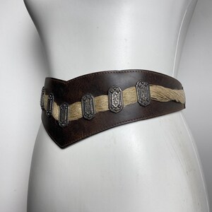 May include: A wide dark brown leather belt featuring a V-shaped front design. The belt is accented with a horizontal strip of light tan fibrous material and a series of rectangular silver-toned metal plates with intricate embossed patterns.