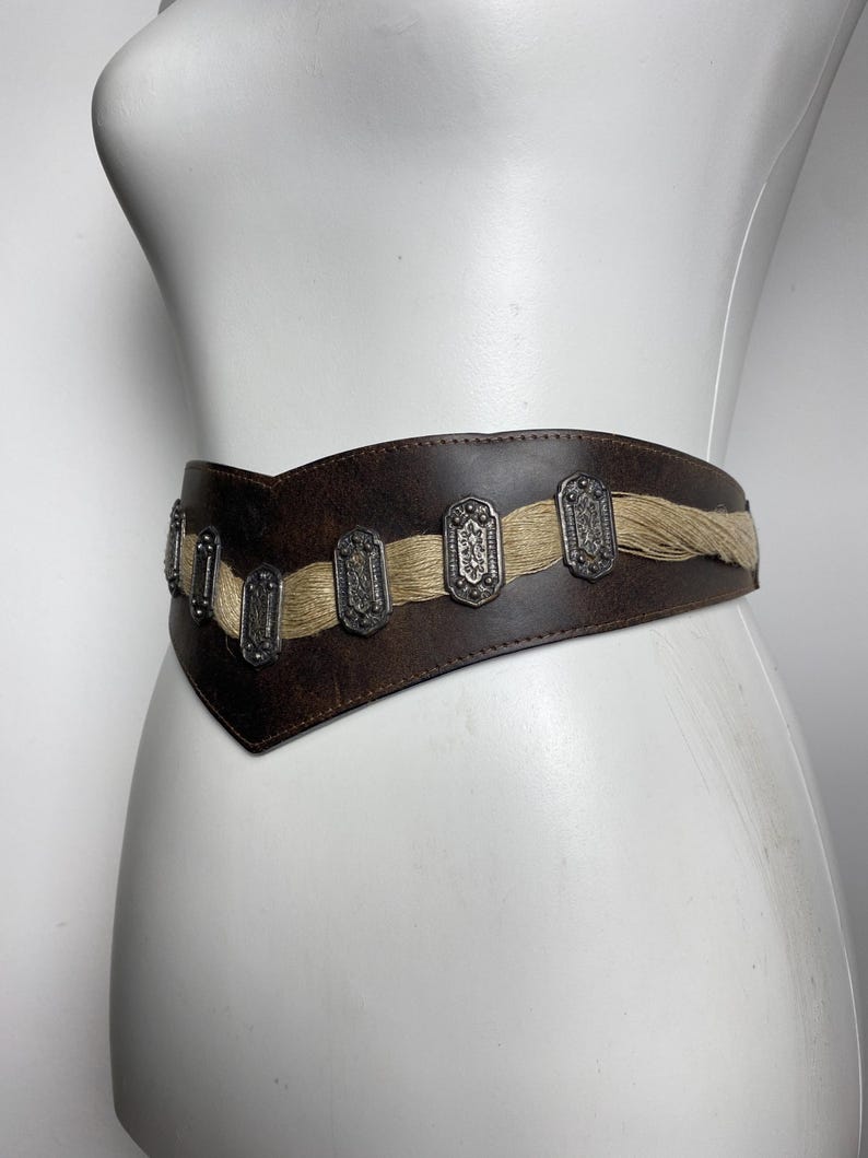 May include: A wide dark brown leather belt featuring a V-shaped front design. The belt is accented with a horizontal strip of light tan fibrous material and a series of rectangular silver-toned metal plates with intricate embossed patterns.