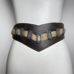 May include: A wide dark brown leather belt featuring a V-shaped front design. The belt is accented with a horizontal band of light tan woven fiber material and seven rectangular silver-toned metal plates with embossed floral patterns.