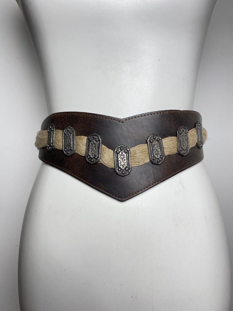 May include: A wide dark brown leather belt featuring a V-shaped front design. The belt is accented with a horizontal band of light tan woven fiber material and seven rectangular silver-toned metal plates with embossed floral patterns.