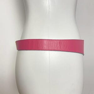 May include: A vibrant pink leather belt is displayed on a white mannequin. The belt is a solid color, with a smooth texture and a rectangular shape. The belt is designed to be worn around the waist.