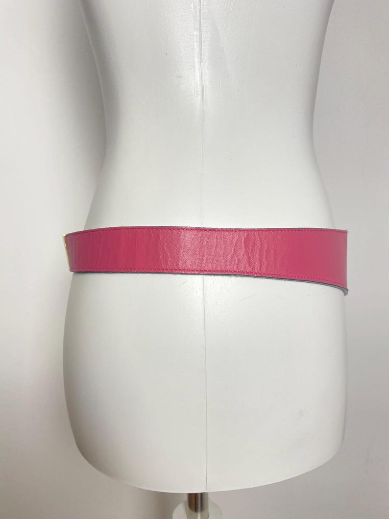 May include: A vibrant pink leather belt is displayed on a white mannequin. The belt is a solid color, with a smooth texture and a rectangular shape. The belt is designed to be worn around the waist.