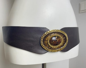 May include: A wide, dark brown leather belt with a large oval buckle. The buckle features a gold chain border around a brown and gold patterned center. The belt is displayed on a white mannequin.