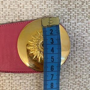 May include: A pink leather belt with a large, round gold-tone buckle. The buckle features a sunburst design. The belt is being measured with a blue measuring tape. The buckle is approximately 3 inches in diameter.