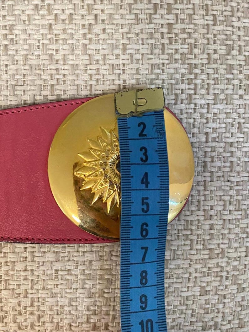 May include: A pink leather belt with a large, round gold-tone buckle. The buckle features a sunburst design. The belt is being measured with a blue measuring tape. The buckle is approximately 3 inches in diameter.