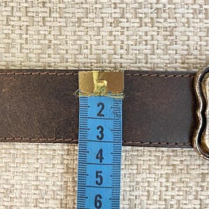 May include: A dark brown leather belt with visible stitching along the edges, positioned next to a blue measuring tape that shows the belt width is approximately 1.5 inches. A metallic buckle is partially visible on the right side of the belt.