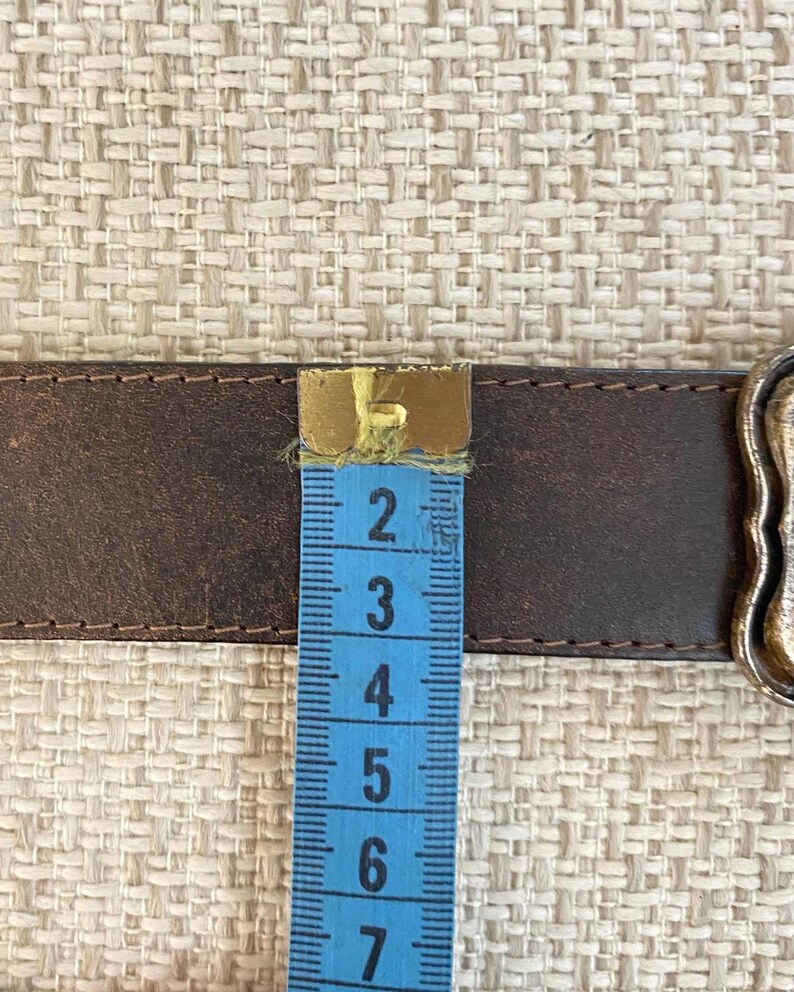 May include: A dark brown leather belt with visible stitching along the edges, positioned next to a blue measuring tape that shows the belt width is approximately 1.5 inches. A metallic buckle is partially visible on the right side of the belt.