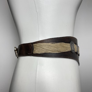 May include: A wide dark brown belt featuring a central horizontal panel of light tan fibrous material. The belt is accented with two rectangular silver-toned metal ornaments with intricate embossed detailing and a silver-toned metal buckle closure.