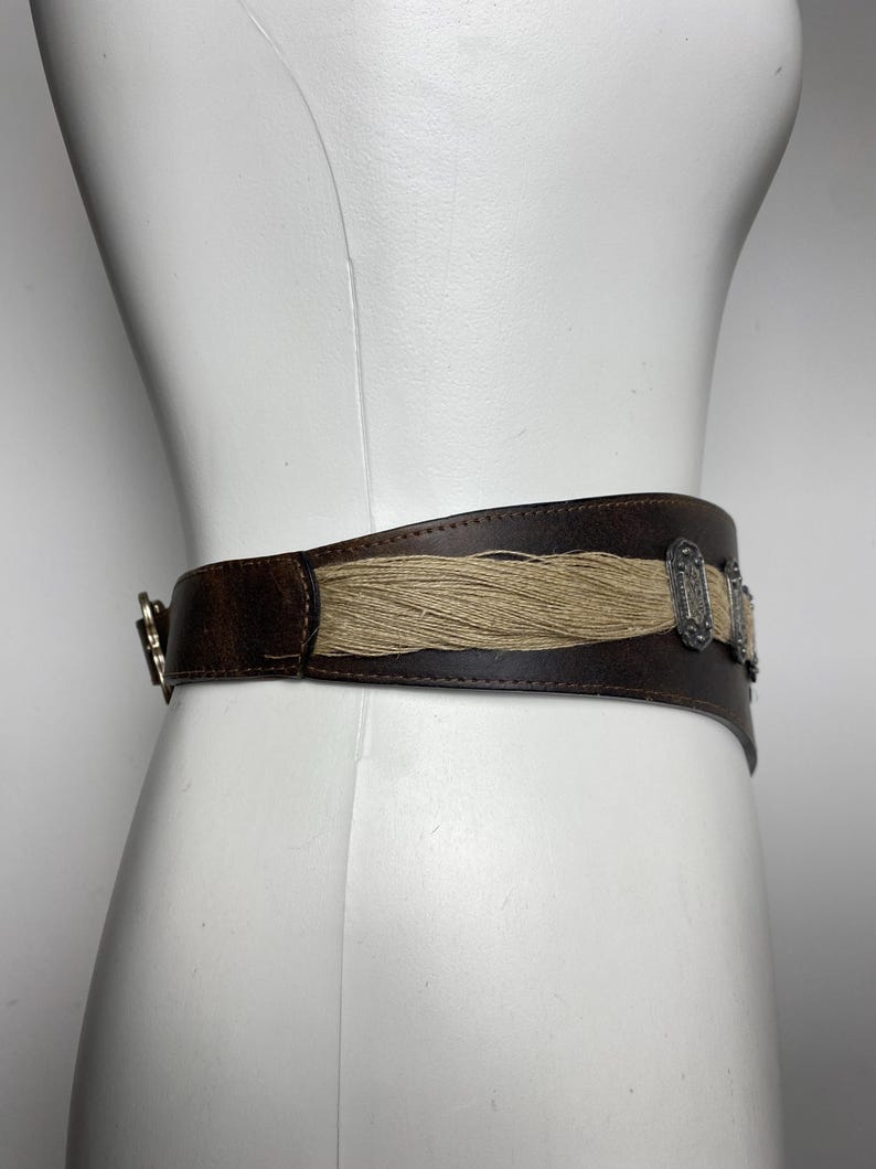 May include: A wide dark brown belt featuring a central horizontal panel of light tan fibrous material. The belt is accented with two rectangular silver-toned metal ornaments with intricate embossed detailing and a silver-toned metal buckle closure.