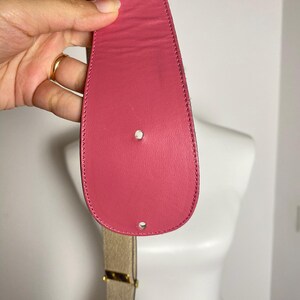 May include: A pink leather belt with a gold-colored buckle. The belt is held up by a hand. The belt's end has two holes.