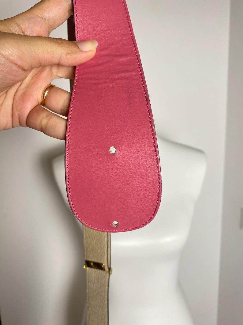 May include: A pink leather belt with a gold-colored buckle. The belt is held up by a hand. The belt's end has two holes.