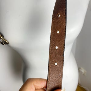 May include: A brown leather belt strap featuring a series of evenly spaced circular adjustment holes. The leather has a visible grain texture and dark edges, with a metallic buckle partially visible on the left side of the frame.