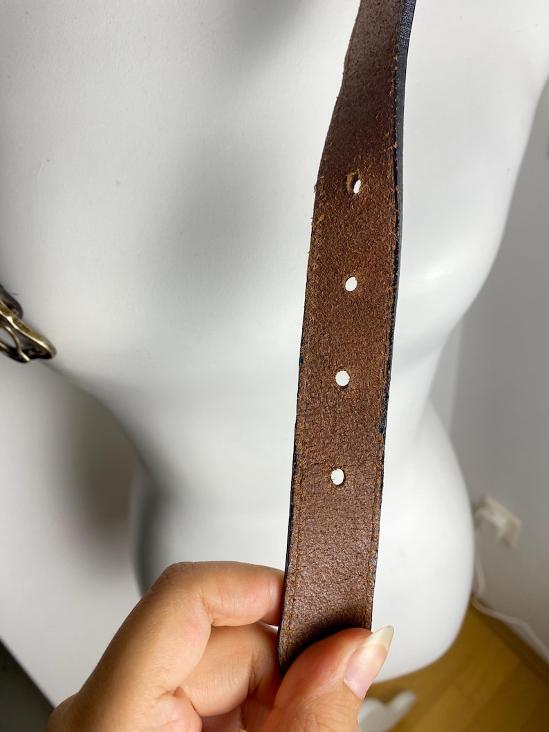 May include: A brown leather belt strap featuring a series of evenly spaced circular adjustment holes. The leather has a visible grain texture and dark edges, with a metallic buckle partially visible on the left side of the frame.