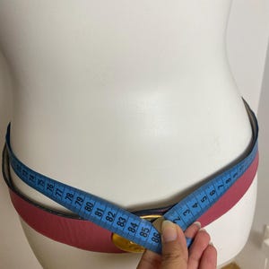 May include: A pink leather belt with a gold-colored buckle is measured with a blue measuring tape. The belt is displayed on a white mannequin. The belt's measurement is approximately 86 cm.