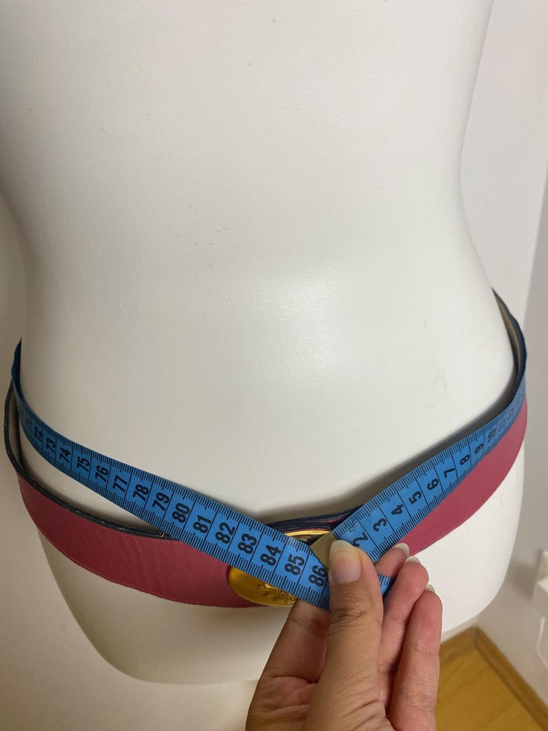May include: A pink leather belt with a gold-colored buckle is measured with a blue measuring tape. The belt is displayed on a white mannequin. The belt's measurement is approximately 86 cm.