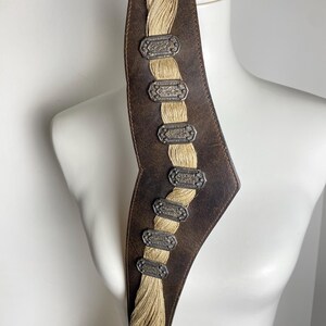 May include: A dark brown leather belt featuring a central vertical panel of braided beige cord. The belt is adorned with seven rectangular, silver-toned metal plaques with intricate embossed detailing, spaced evenly along the braided cord section.