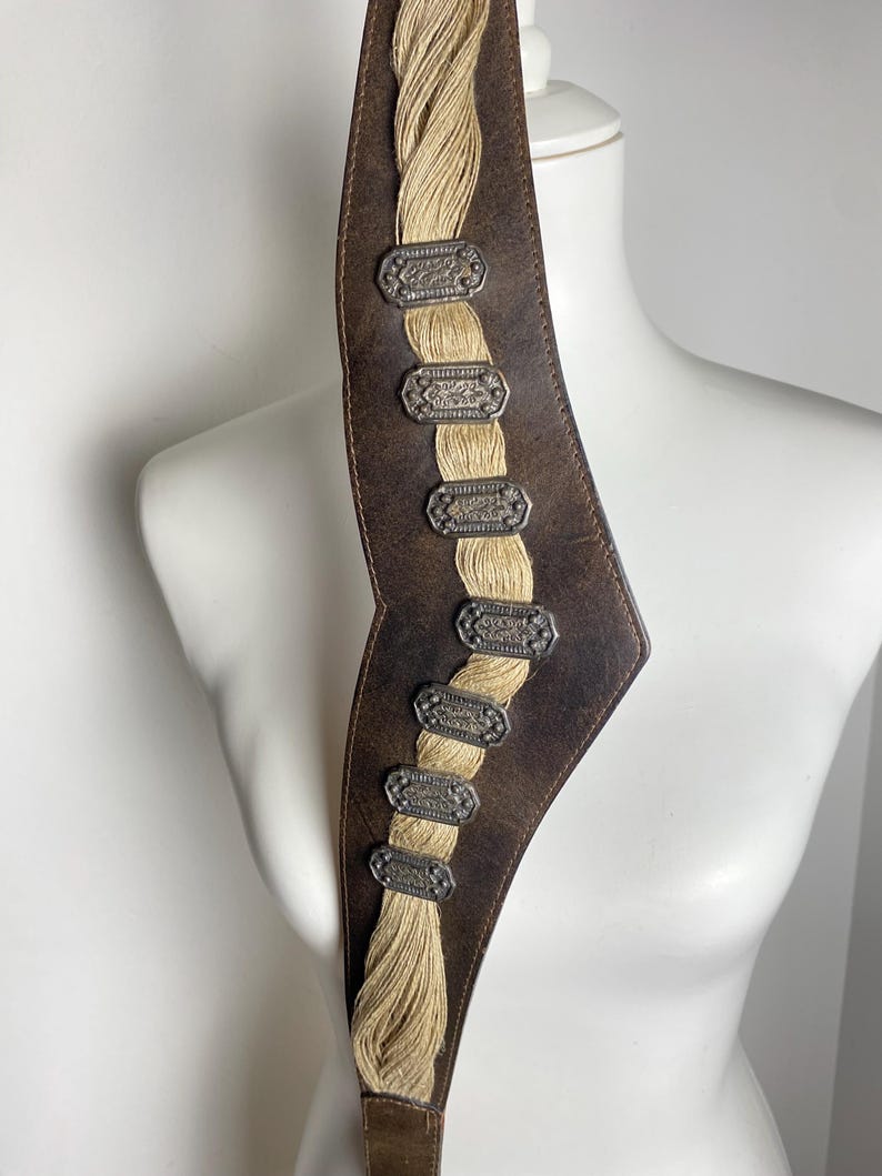 May include: A dark brown leather belt featuring a central vertical panel of braided beige cord. The belt is adorned with seven rectangular, silver-toned metal plaques with intricate embossed detailing, spaced evenly along the braided cord section.