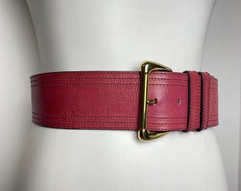 May include: A wide, red leather belt with a gold-colored buckle. The belt is wrapped around a white mannequin. The belt has a stitched design and is approximately 3 inches wide.