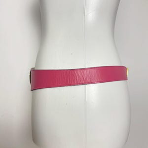 May include: A bright pink leather belt with a gold-colored buckle is displayed on a white mannequin. The belt is wrapped around the mannequin's waist, showcasing its design and color. The belt's width is approximately 1.5 inches.
