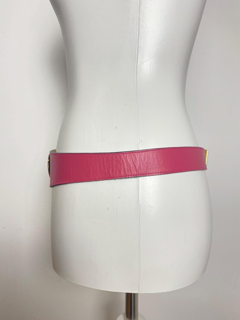 May include: A bright pink leather belt with a gold-colored buckle is displayed on a white mannequin. The belt is wrapped around the mannequin's waist, showcasing its design and color. The belt's width is approximately 1.5 inches.