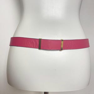 May include: A hot pink leather belt with gold-colored metal hardware. The belt is displayed on a white mannequin. The belt is a classic accessory.