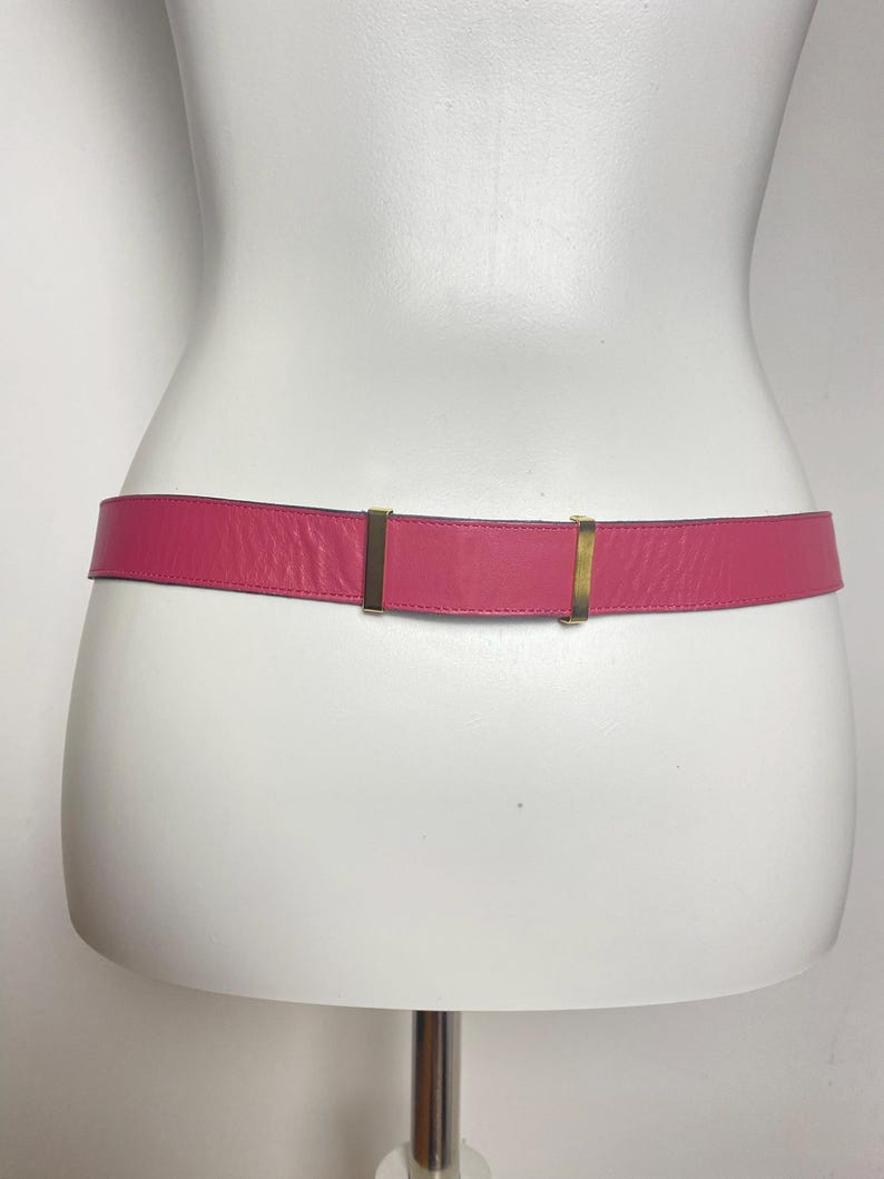 May include: A hot pink leather belt with gold-colored metal hardware. The belt is displayed on a white mannequin. The belt is a classic accessory.