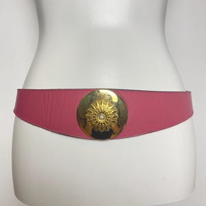 May include: A pink leather belt with a gold-tone circular buckle. The buckle features a sunburst design with a central rhinestone. The belt is displayed on a white mannequin.