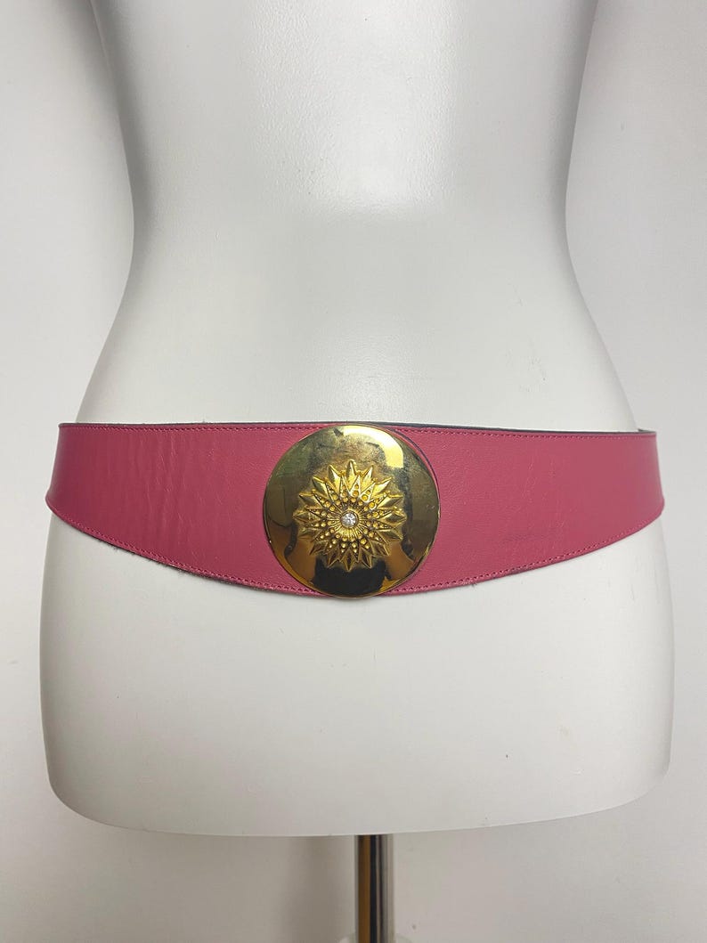 May include: A pink leather belt with a gold-tone circular buckle. The buckle features a sunburst design with a central rhinestone. The belt is displayed on a white mannequin.