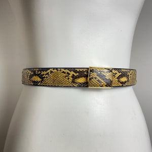 May include: A snakeskin belt with a gold-colored buckle is displayed on a white mannequin. The belt features a pattern of yellow, brown, and black scales. The buckle is rectangular and appears to be made of metal.