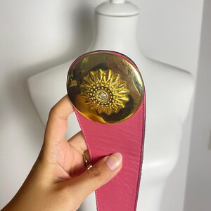 May include: A pink leather belt with a large gold-tone buckle. The buckle features a sunburst design with a central crystal. The belt is held up by a hand.