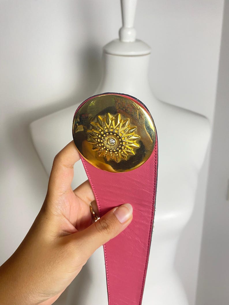 May include: A pink leather belt with a large gold-tone buckle. The buckle features a sunburst design with a central crystal. The belt is held up by a hand.