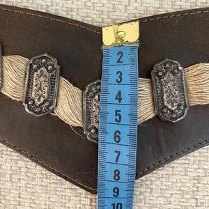 May include: A dark brown leather belt featuring three rectangular silver-toned metal ornaments with intricate embossed patterns, accented by beige fiber fringe. A blue measuring tape shows the belt width is approximately 3.5 inches.
