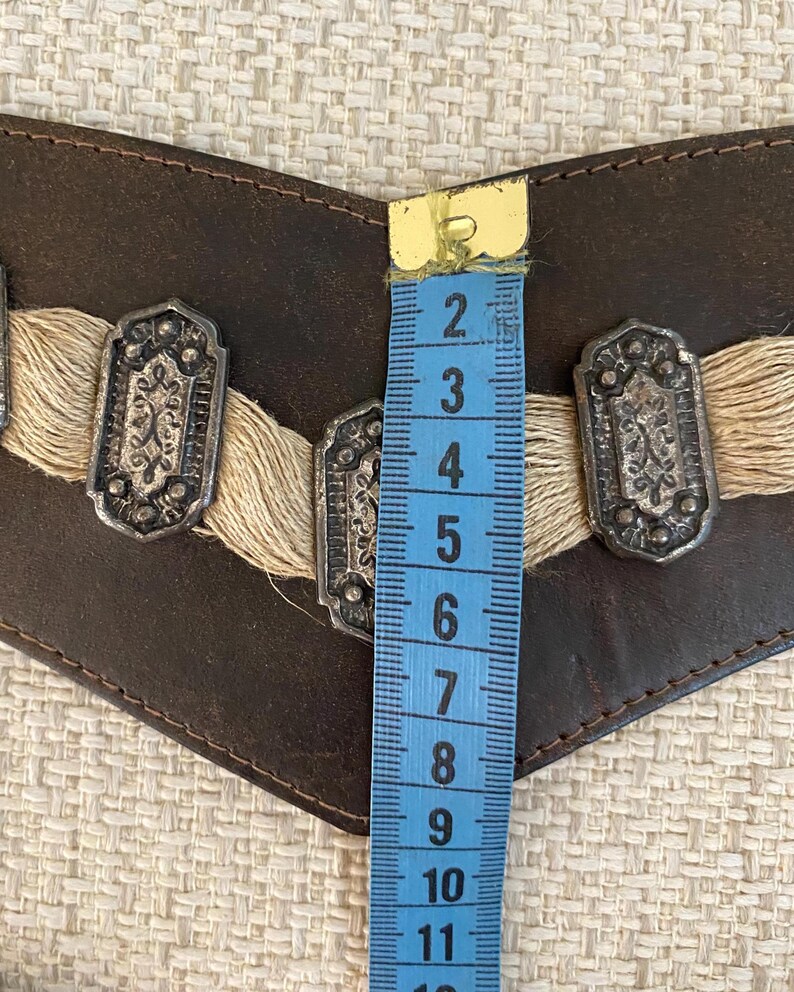 May include: A dark brown leather belt featuring three rectangular silver-toned metal ornaments with intricate embossed patterns, accented by beige fiber fringe. A blue measuring tape shows the belt width is approximately 3.5 inches.