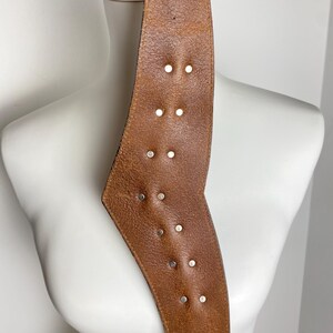 May include: A wide, tan-colored leather belt featuring a distinct angular shape and a series of small, circular silver-toned metal studs arranged in pairs along the center. The leather has a visible grain texture and a smooth, finished edge.