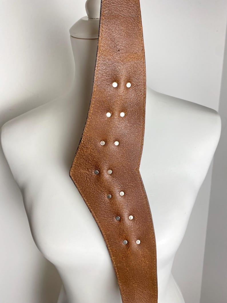May include: A wide, tan-colored leather belt featuring a distinct angular shape and a series of small, circular silver-toned metal studs arranged in pairs along the center. The leather has a visible grain texture and a smooth, finished edge.