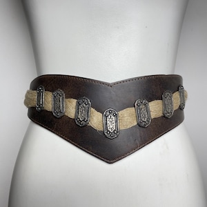 May include: A wide, dark brown leather belt featuring a V-shaped front design. The belt is accented with a horizontal band of light tan, textured fiber material and seven rectangular, silver-toned metal plates with intricate embossed patterns.