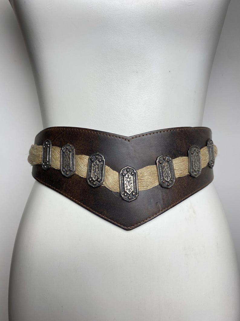 May include: A wide, dark brown leather belt featuring a V-shaped front design. The belt is accented with a horizontal band of light tan, textured fiber material and seven rectangular, silver-toned metal plates with intricate embossed patterns.