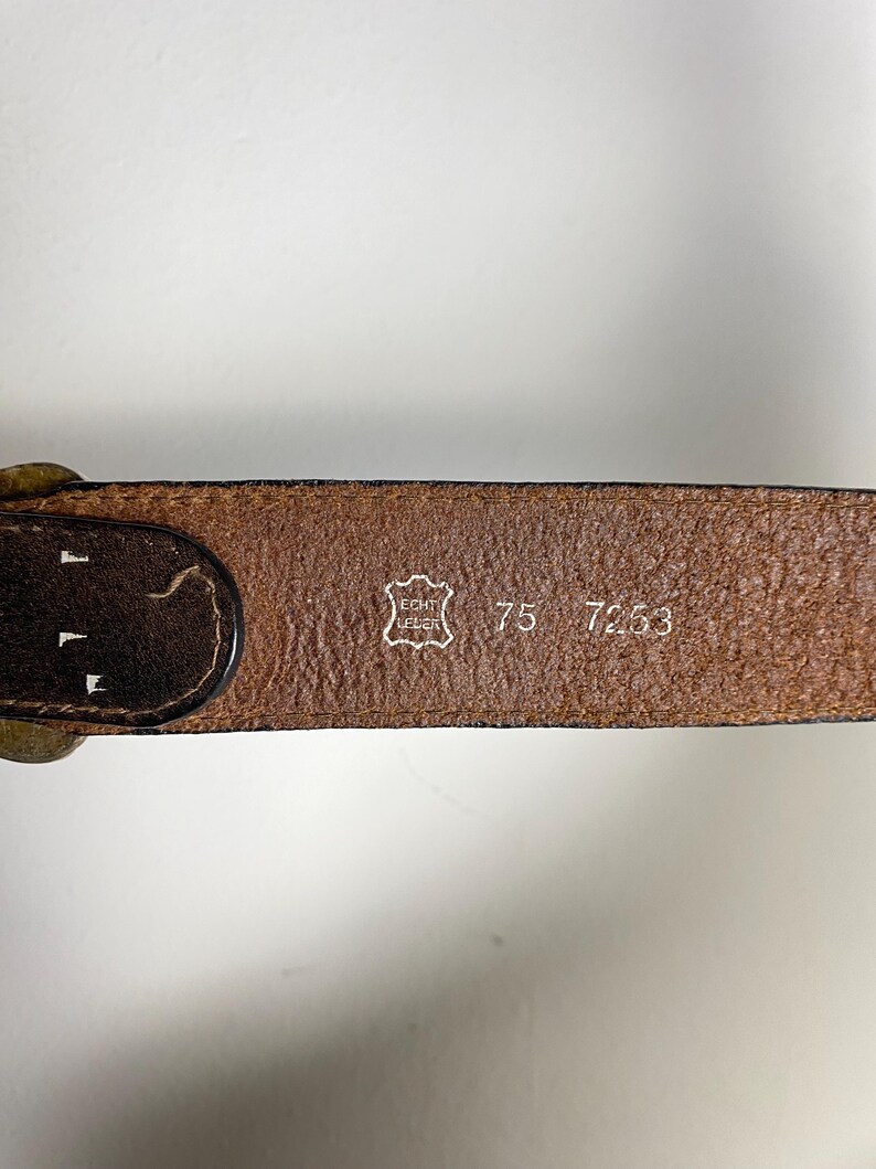 May include: A brown leather belt featuring a stamped emblem with the text ECHT LEDER, the number 75, and the number 7253. The belt has a dark brown edge and a metallic buckle visible on the left side, showing a textured surface and clear markings.