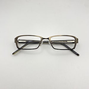 Könnte beinhalten: Rechteckige Brille mit bronzefarbenem Rahmen und klaren Gläsern. Die Brille hat ein modernes Design mit schmalem Steg und Bügeln. Die Bügel sind dunkel und die Gläser sind klar.