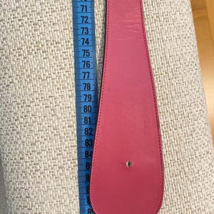 May include: A bright pink leather belt with a rounded end, shown with a measuring tape. The belt is approximately 33 inches long. The belt has two holes for the buckle. The background is a textured, light-colored fabric.