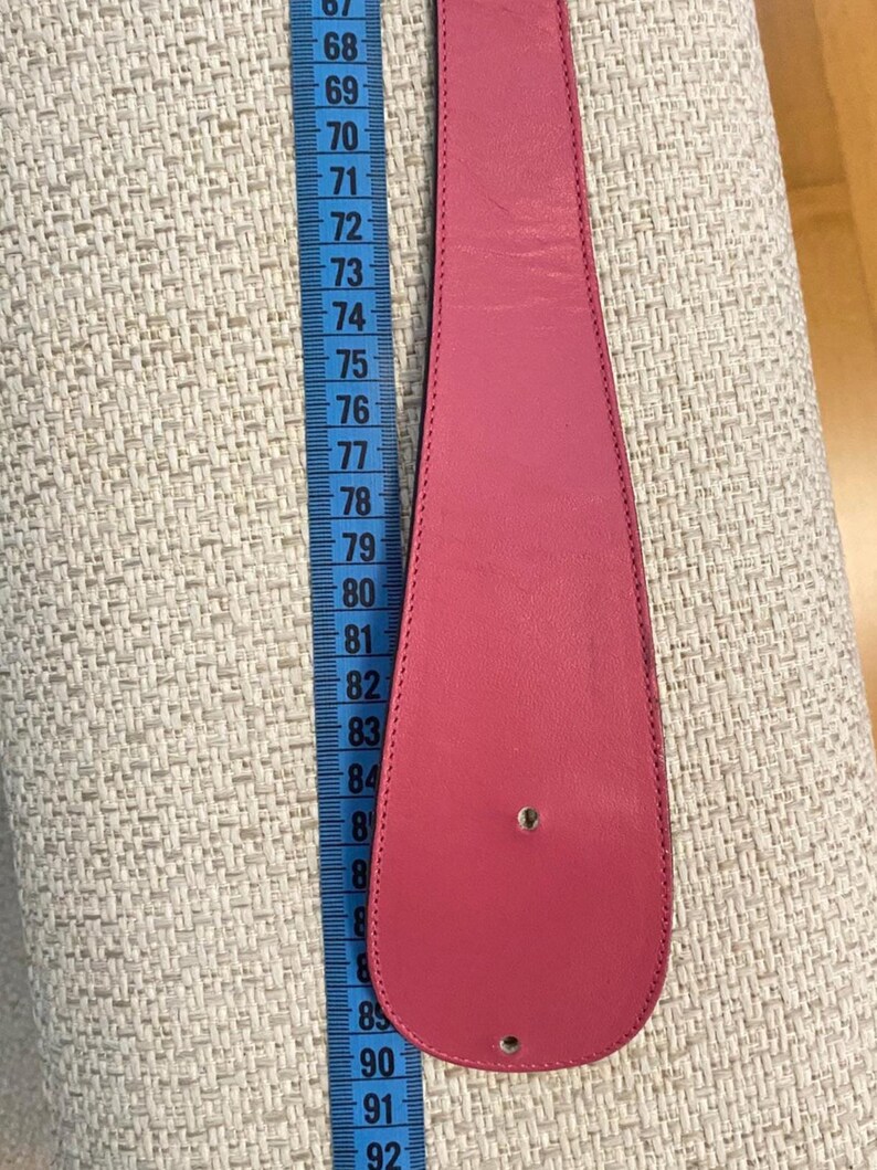 May include: A bright pink leather belt with a rounded end, shown with a measuring tape. The belt is approximately 33 inches long. The belt has two holes for the buckle. The background is a textured, light-colored fabric.